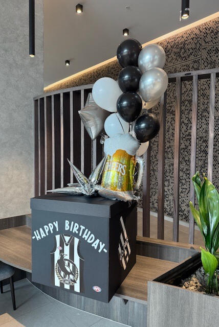 Collingwood AFL Birthday Box with Balloons A black Collingwood-themed birthday gift box with helium balloons in black, white and silver. Features a footy jersey design, silver stars and a large beer mug balloon with "Cheers".
